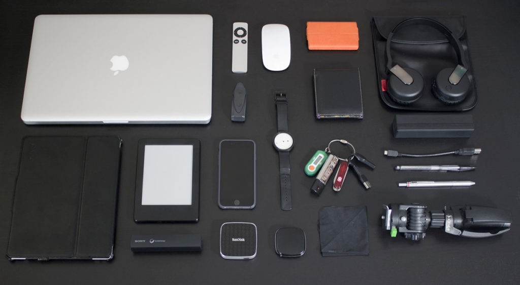 A table full of every day carry items like a MacBook, headphones, iPhone, watch, tripod, mouse, iPad, and pens.